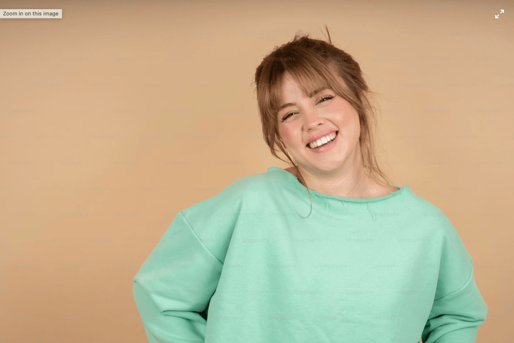 Woman with reddish-brown hair and bangs smiling warmly, wearing a mint green oversized sweater against a tan background