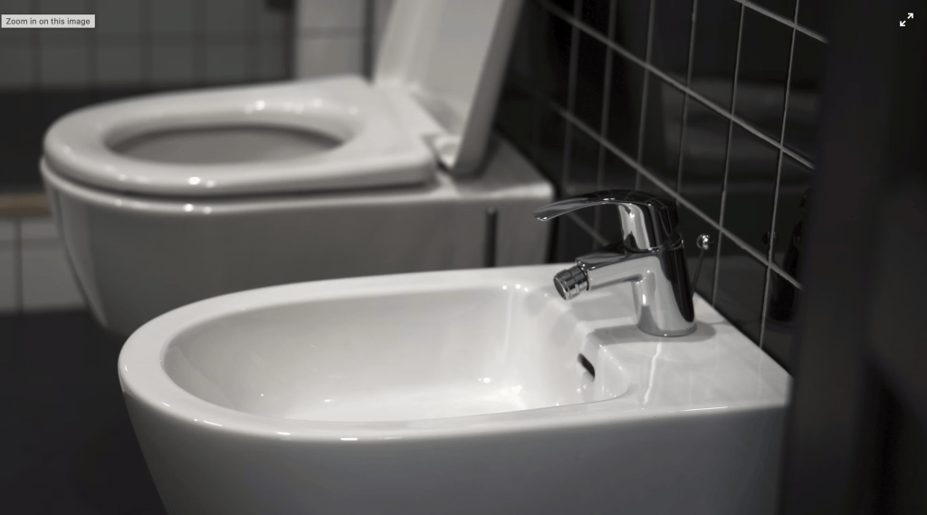 A white bidet next to a white toilet in a bathroom with black tiles.
