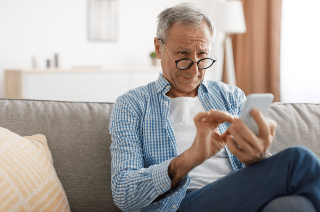 Gray-haired man in glasses and a blue checkered shirt sits on a couch, squinting slightly as he taps the screen of a smartphone