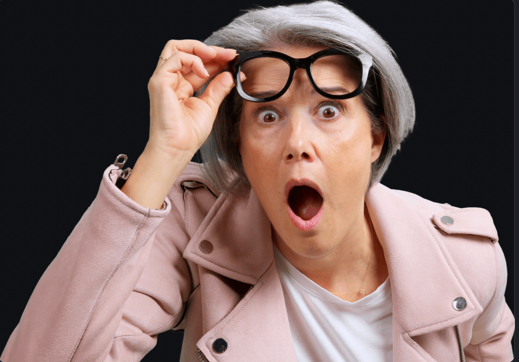 Older woman with gray hair lifting her black-framed glasses and looking shocked, mouth open, wearing a pink jacket against a black background