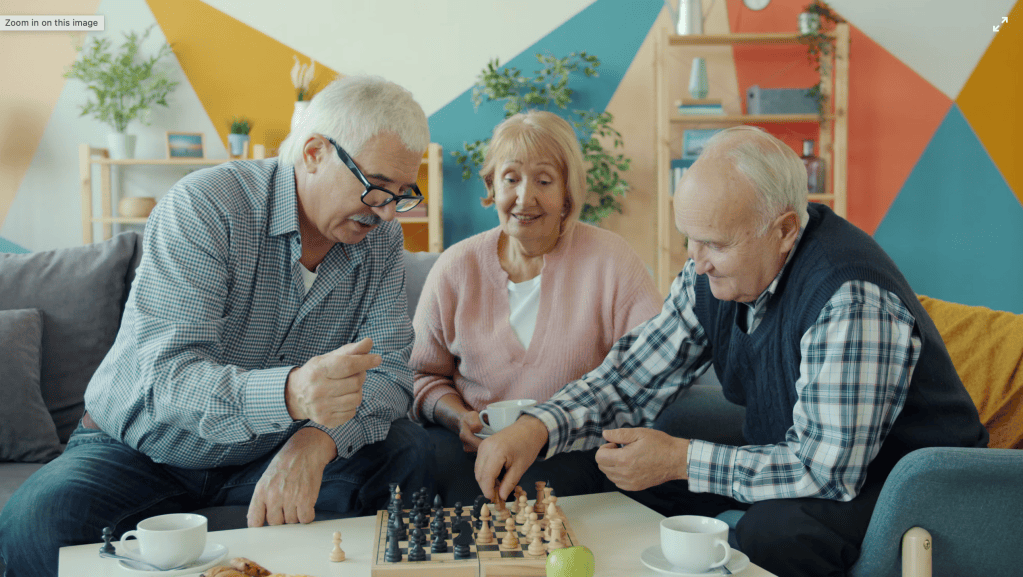 Three older adults sitting on a couch playing chess together while sharing coffee in a colorful living room