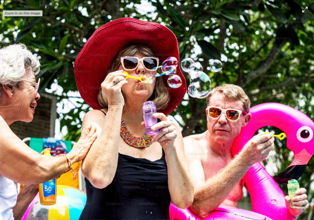Older adults playing with bubble wands and sunscreen at a colorful pool party, one wearing a wide red hat and another holding an inflatable flamingo
