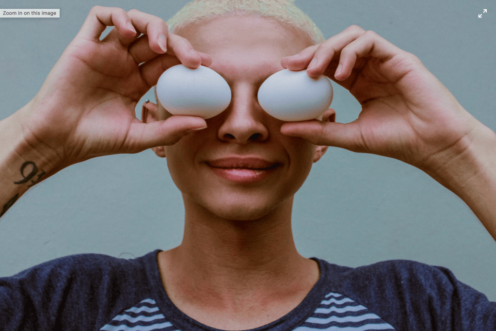 Person with short bleached hair smiling and holding two white eggs over their eyes like goggles